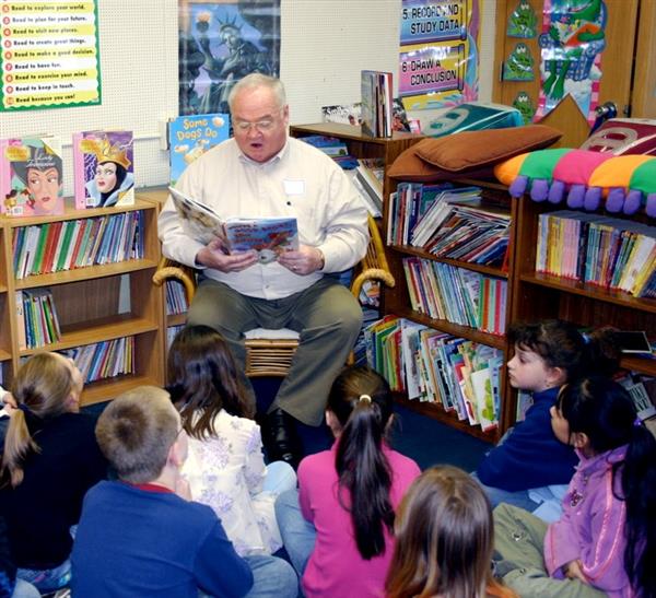 Werner Kleemann is seated reading a book to elementary students